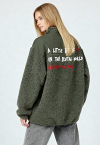 Veste en polaire vert olive avec une surface texturée, comportant un texte imprimé blanc et rouge dans le dos. Coupe ample, col montant et manches longues.