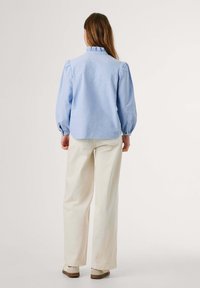 Lichtblauwe blouse met lange mouwen, versierd met een rimpelcol en gathered mouwen, gecombineerd met crèmekleurige, wijde broekspijpen en lichte schoenen.