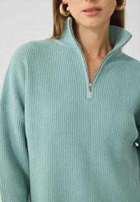 Jasnoniebieski sweter prążkowany z kołnierzykiem na pół zamku i miękką teksturą, uzupełniony przez dużą okrągłą złotą biżuterię w uszach.