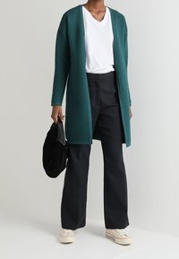 Tealribbat kavaj över en vit V-ringad T-shirt, ihop med svarta vida byxor och krämfärgade sneakers. Håller en svart väska.