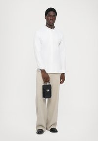 Witte blouse met lange mouwen, beige wijde broek, houdt een kleine zwarte tas met zilveren accenten vast, staande tegen een neutrale achtergrond.