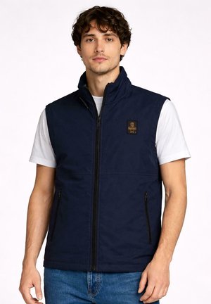 Uomo che indossa un gilet senza maniche blu navy con zip sopra una camicia bianca a maniche corte e jeans blu, in piedi davanti a uno sfondo semplice.