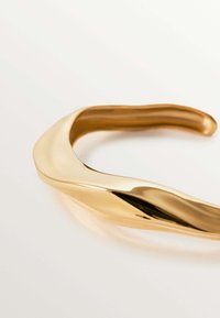 Collar de metal en tono dorado, diseño suave y ondulado con superficie reflectante y una forma fluida, sin cierres o adornos visibles.