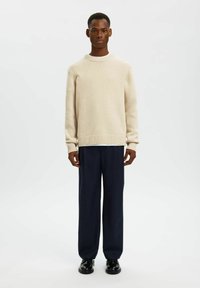 Beige stickad tröja med rund halsringning och ribbade muddar och fåll, ihop med mörkblå vida byxor och svarta skor. Slät textur, avslappnad passform.