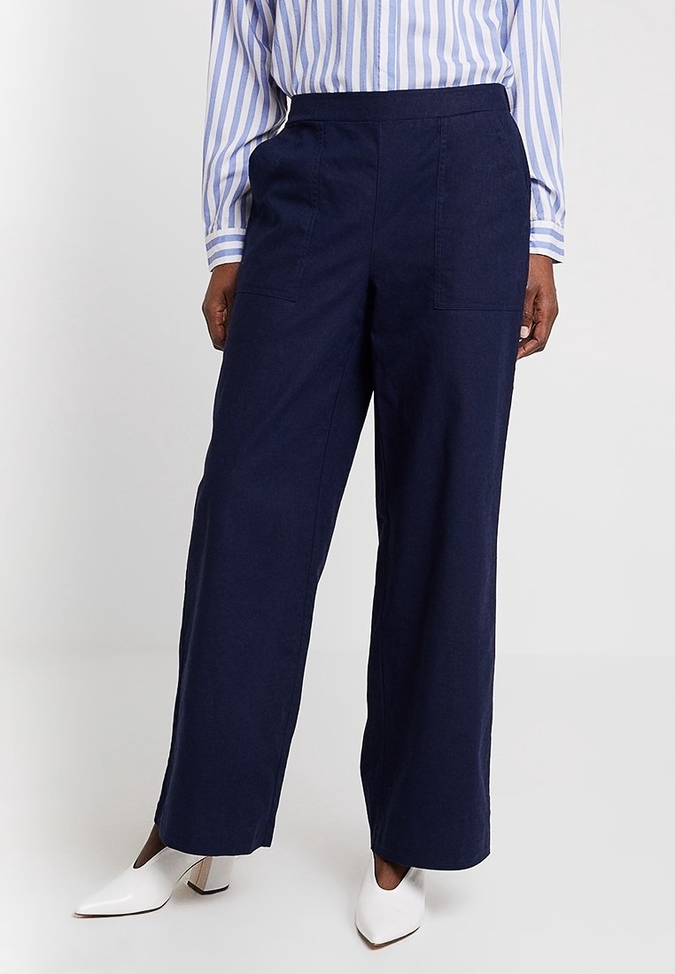 Personne portant un pantalon pattes d'éléphant bleu marine, une chemise à rayures bleues et blanches à boutons, et des chaussures blanches à talons, se tenant contre un fond uni.