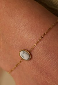 Gold chain bracelet featuring an oval white stone with gray marbling, textured metal detailing around the stone, resting on skin.