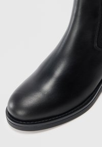 Gros plan sur la pointe d'une botte en cuir noir lisse, de forme arrondie, avec une semelle cousue sur un fond blanc uni.