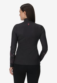 Maglietta nera a maniche lunghe con colletto alto, caratterizzata da un logo di ragno rosso sul retro e motivi floreali colorati sulle maniche.