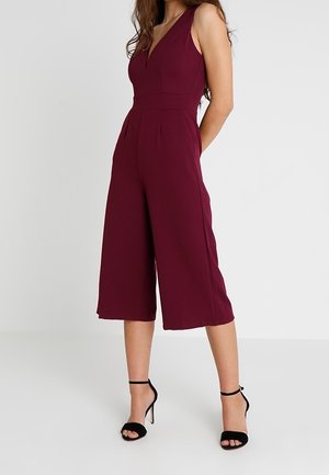 Bordeauxrode jumpsuit met een v-hals en mouwloos ontwerp. Wijd uitgesneden culottes en getailleerde naden zorgen voor een gestructureerde uitstraling. Gecombineerd met zwarte sandalen met hak.