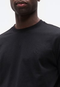 Άνδρας που φοράει μαύρο T-shirt με στρογγυλή λαιμόκοψη και μικρό, διακριτικό λογότυπο "ck" κεντημένο στο αριστερό στήθος.