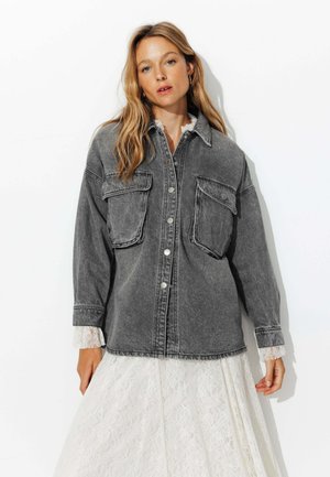 Femme portant une veste en denim surdimensionnée noire délavée sur une robe en dentelle blanche, se tenant devant un fond blanc uni.