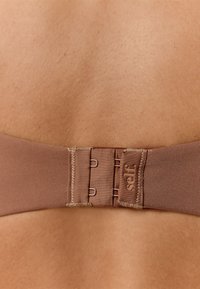 Pruun kangast rinnahoidja pael konksude ja silmadega reguleerimiseks. Omadusteks on tekstuuriga pind ja õmblusdetailid, küljesilt "self.".