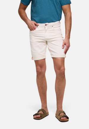 Hombre con pantalones cortos color crema enrollados, camisa de manga corta verde azulado y sandalias marrones con dos hebillas, de pie contra un fondo blanco.