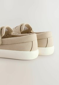 Beiga mockakskor med en låg design, vita gummisulor och dubbla kardborreband. Har en mjuk foder och synliga sömnadsdetaljer.