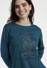 Mujer joven sonriendo, vestida con una camiseta de manga larga en color teal con un Mickey Mouse bordado en dorado y el texto "Haz lo que amas, ama lo que haces."