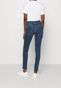 Jeans de denim azul ajustados con una cintura de altura media, un desgaste sutil y dos bolsillos traseros, combinados con una camiseta blanca sencilla y zapatillas blancas.