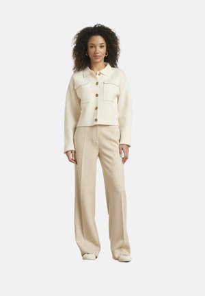 Vrouw staand, gekleed in een lichtbeige jasje met knopen, twee borstzakken en bijpassende wijde pijpen en witte schoenen.