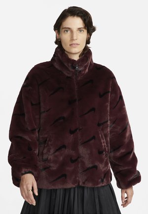 Persone che indossa una giacca in pile bordeaux con motivi neri a forma di swoosh Nike e una gonna nera a pieghe su uno sfondo bianco.