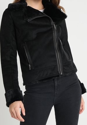 Vrouw draagt een zwart jas met imitatiebontvoering, een asymmetrische ritssluiting en ritssluitingen aan de zijkanten, gecombineerd met zwarte jeans.