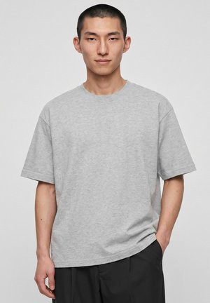 Jeune homme asiatique aux cheveux courts portant un t-shirt uni gris clair à manches courtes et un pantalon noir, debout avec une main dans la poche.