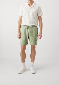 Chemise blanche texturée à manches courtes avec une poche poitrine, associée à un short léger vert pâle avec cordon de serrage, comprenant une poche latérale et un patch logo.