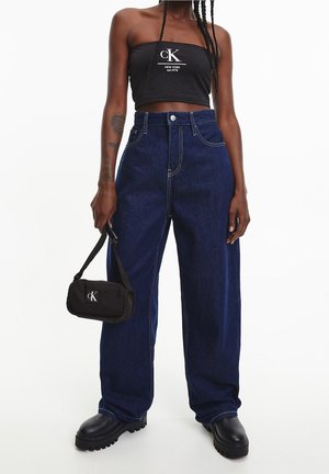 Femme portant un haut tube noir CK, jean taille haute à jambes larges bleu foncé, bottes noires épaisses, tenant un petit sac à main noir CK.