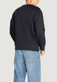 Felpa nera con motivo logo tono su tono su tutta la superficie, maniche a costine e vita a costine. Abbinata a jeans in denim azzurri.