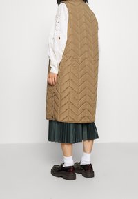 Hnědá prošívaná vesta s vzorem chevron, spárovaná s skládacími tmavozelenými sukněmi, nošena s černými platformovými botami a bílými ponožkami.