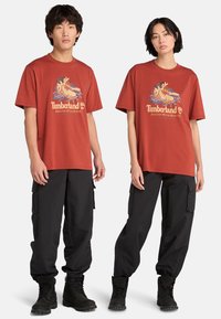 Rustrøde bomulds t-shirts med grafisk print, parret med sorte cargo bukser og sorte snørebogs støvler. Casual fit, korte ærmer.