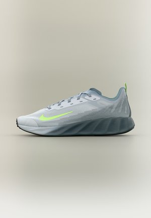 Zapatilla deportiva gris claro con el logo de Nike en verde neón, parte superior de malla y suela gruesa y texturizada en gris oscuro sobre un fondo neutro.