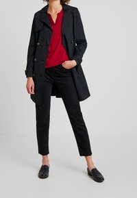 Kvinde iført en sort trench coat, rød bluse, sorte cropped jeans og sorte loafers, der står med den ene hånd i lommen foran en ensfarvet baggrund.