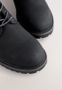 Čierne kožené topánky s textúrovaným povrchom, okrúhlou špičkou, sivými šnúrkami a hrubou gumovou podrážkou. Detaily zahŕňajú viditeľné švy a vzor dezénu.