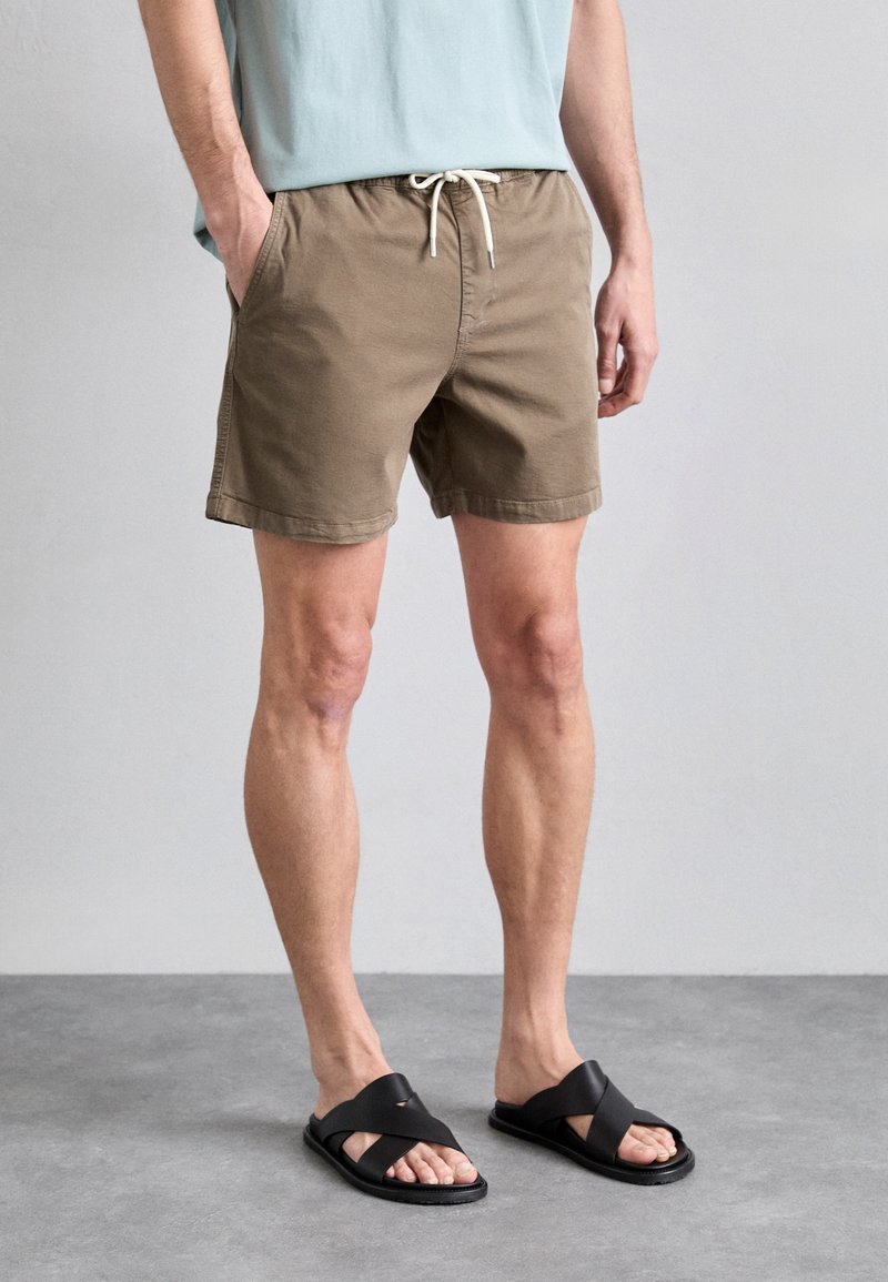 Khaki katoenen shorts met een elastische tailleband en trekkoord, gecombineerd met zwarte sandalen met kruisbanden. Korte lengte en casual pasvorm.