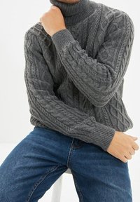 Grauer Rollkragenpullover aus Zopfstrick mit gerippten Bündchen und Halsabschluss, über blauen Jeans getragen und zeigt ein strukturiertes Design.