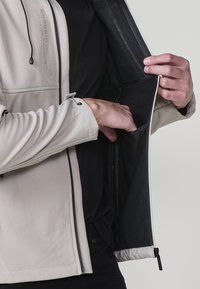 Béžová bunda so zipsom a vreckami; hladká tkanina s jemným leskom. Vnútorné podšívka je čierna a obsahuje minimálne švy a detaily z kovania.