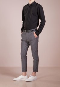 Mann trägt ein schwarzes Hemd mit Knöpfen, graue maßgeschneiderte Hose mit Gürtel, weiße Turnschuhe, steht mit den Händen in den Taschen vor beigem Hintergrund.