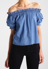 Blusa in cotone leggera blu chiaro con spalline scoperte, collettore elastico arricciato al collo e maniche a sbuffo. Tessuto liscio, vestibilità casual.