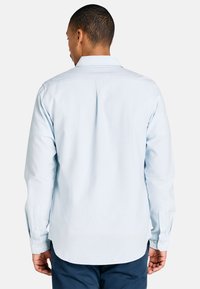 Uomo visto di spalle che indossa una camicia a maniche lunghe azzurro chiaro e pantaloni blu scuro, su uno sfondo bianco semplice.
