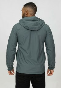 Veste grise résistante à l'eau avec capuche, ayant une texture lisse et des poignets réglables, vue de dos.