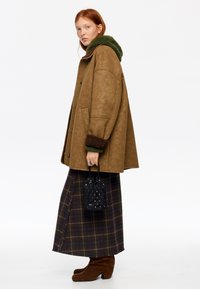 Chaqueta marrón de cuero sintético con cuello de piel sintética verde, combinada con una falda a cuadros y botines de ante marrón. Bolso negro con tachuelas en la mano.