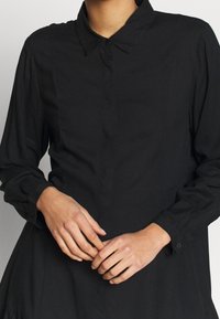 Camicia nera con colletto a punta, maniche lunghe e tessuto leggermente strutturato; presenta una patta frontale e polsini aderenti.