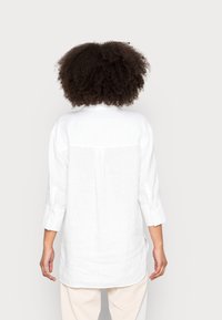 Chemise en lin blanc à coupe décontractée, manches longues retroussées jusqu'aux coudes, avec une couture de yoke au dos. Comprend un ourlet droit et une texture subtile.