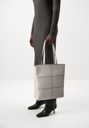 Tasche aus hellgrauem Leder mit einem quadratischen Muster, ausgestattet mit zwei Schulterriemen. Getragen zu einem dunklen, gerippten Kleid und schwarzen, spitz zulaufenden Stiefeln.