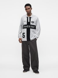 Jeune homme portant un polo à manches longues avec une croix noire et du texte, un pantalon cargo ample et foncé, et des baskets noires, debout avec une main dans la poche.