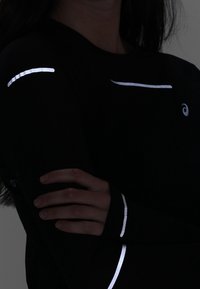 Camiseta negra de manga larga deportiva con acentos reflectantes en los hombros y las mangas. Tejido texturizado con un diseño ajustado.
