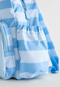 Le sac à dos rayé bleu présente un tissu extérieur texturé, une fermeture zippée et un détail de poche froncée. Les bretelles ajustables bleues complètent le design.