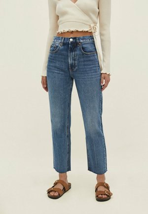 Vrouw draagt blauwe high-waist rechte jeans, beige wikkeltrui met geruite randen en bruine sandaal met gesp op lichte achtergrond.
