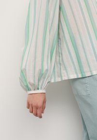 Blouse rayée avec des teintes vertes et beiges, manches longues avec des poignets élastiques et une coupe décontractée. Le tissu semble léger et texturé.