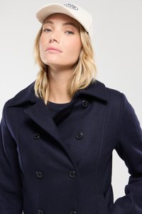 Manteau bleu marine croisé en laine avec une finition texturée et des boutons noirs ; associé à une casquette de couleur claire arborant un logo.