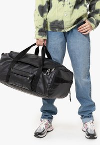 Czarna torba sportowa wykonana z trwałego nylonu, z dwiema górnymi uchwytami, przednią kieszenią na zamek błyskawiczny oraz regulowanym paskiem. Charakteryzuje się smukłym, zaokrąglonym kształtem.